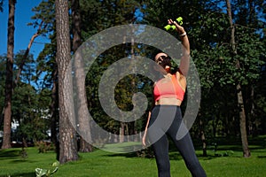 Young sporty woman raising her arm with dumbbells up ad looking at it standing in a city park