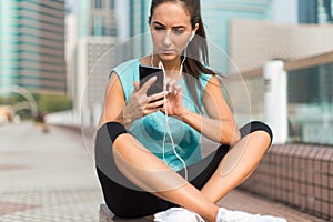 Young sporty woman resting after exercising using her smartphone and listening to music in earphones. Athlete runner in