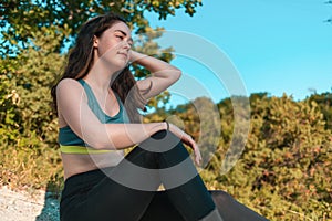 Young sporty woman resting, sitting on the grass. Forest in the background. Sports, fitness and healthy lifestyle tracking concept