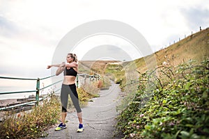 Young sporty woman runner in black activewear standing outside in nature, stretching.