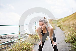 Young sporty woman runner in black activewear standing outside by the seaside, resting.