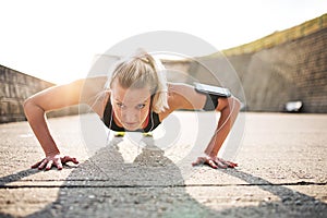 Young sporty woman runner with smartphone doing push-ups outside.