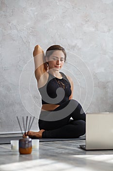 Young sporty woman watching online yoga lessons on laptop. Beautiful girl practicing cow face pose, gomukhasana. Yoga