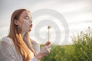 Young spring fashion woman blowing dandelion in spring garden. Springtime. Trendy girl at sunset in spring landscape background.