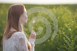 Young spring fashion woman blowing dandelion in spring garden. Springtime. Trendy girl at sunset in spring landscape background.