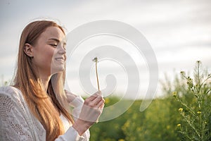 Young spring fashion woman blowing dandelion in spring garden. Springtime. Trendy girl at sunset in spring landscape background.