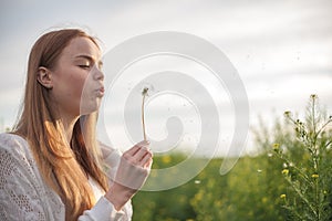 Young spring fashion woman blowing dandelion in spring garden. Springtime. Trendy girl at sunset in spring landscape background.