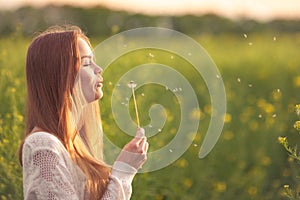 Young spring fashion woman blowing dandelion in spring garden. Springtime. Trendy girl at sunset in spring landscape background.