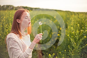 Young spring fashion woman blowing dandelion in spring garden. Springtime. Trendy girl at sunset in spring landscape background