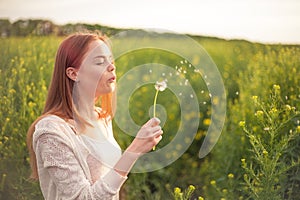 Young spring fashion woman blowing dandelion in spring garden. Springtime. Trendy girl at sunset in spring landscape background