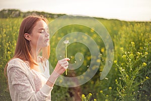 Young spring fashion woman blowing dandelion in spring garden. Springtime. Trendy girl at sunset in spring landscape background