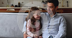 Young stepfather talks to little daughter seated on sofa