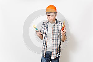 Young strange handsome man in casual clothes and protective orange hardhat holding toy hammer and saw isolated on white