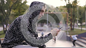 Young streetstyle man in hoodie using his smartphone in hood, Sitting on the fence of skatepark