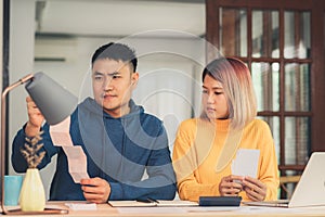 Young stressed asian couple managing finances, reviewing their bank accounts using laptop computer and calculator at modern home.