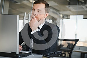 Young stressed handsome businessman working at desk in modern office shouting at laptop screen and being angry about