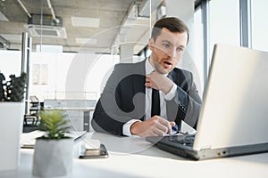 Young stressed handsome businessman working at desk in modern office shouting at laptop screen and being angry about