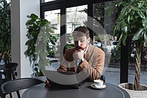 Young stressed nervous annoyed businessman sitting in cafeteria restaurant drinking coffee reluctantly looking at laptop computer