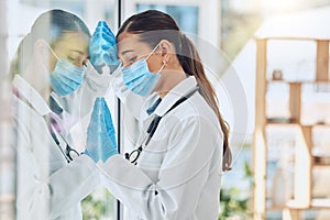 Young stressed and overworked doctor wearing mask and gloves while standing at a window in a hospital or clinic. One