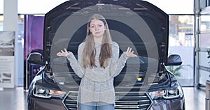 Young stressed woman standing next to broken car with open hood and holding head with hands. Upset Caucasian brunette