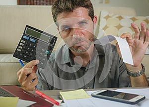 Young stressed and worried man at home living room using calculator and laptop doing domestic accounting paperwork feeling