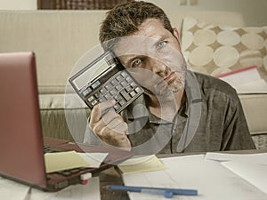 Young stressed and worried man at home living room using calculator and laptop doing domestic accounting paperwork feeling