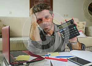 Young stressed and worried man at home living room using calculator and laptop doing domestic accounting paperwork feeling
