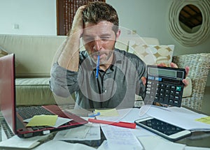 Young stressed and worried man at home living room using calculator and laptop doing domestic accounting paperwork feeling