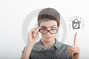 Young strict irritated business woman in glasses pointing index finger up on drawn alarm clock on the white background. Time is