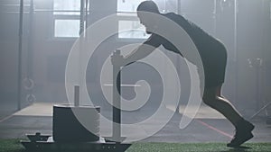 Young strong athletic caucasian man exercising with heavy weight training sled towards camera in large gym slow motion.