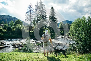 Young strong man stays near mountains river with dog and looking for the view