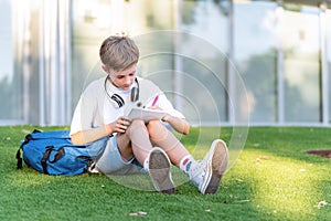 Young student boy doing homework in notebook while sitting on the grass outdoors.