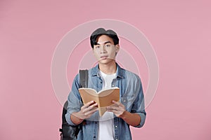 A young student carrying a backpack and holding an open book reading,  on a pink background