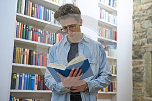 Young student with curls focussed on reading a book and standing  next to a bookshelf photo