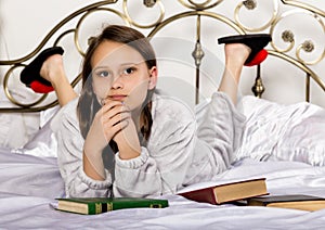 Young student girl reads a book while lying on a bed doing homework