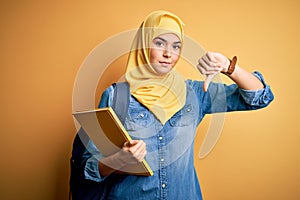 Young student girl wearing muslim hijab and backpack holding book over yellow background with angry face, negative sign showing