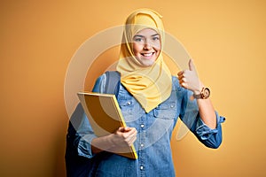 Young student girl wearing muslim hijab and backpack holding book over yellow background happy with big smile doing ok sign, thumb