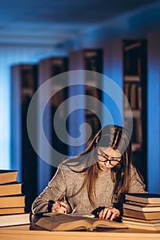 Young student in glasses preparing for the exam. Girl in the evening sits at a table in the library with a pile of books