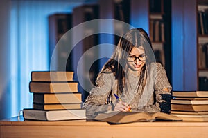 Young student in glasses preparing for the exam. Girl in the evening sits at a table in the library with a pile of books