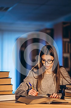Young student in glasses preparing for the exam. Girl in the evening sits at a table in the library with a pile of books