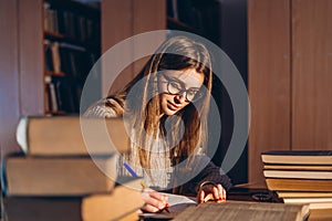 Young student in glasses preparing for the exam. Girl in the evening sits at a table in the library with a pile of books