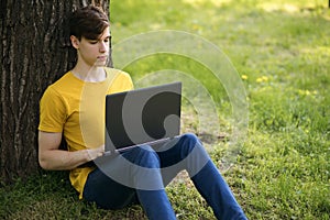 A young student guy sits on the lawn and holds a laptop in his hands