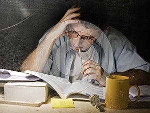 Young student at home desk reading biting pen studying at night with pile of books and coffee