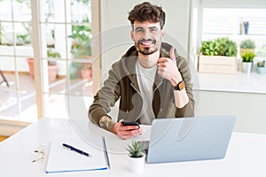 Young student man using smartphone and studying using laptop happy with big smile doing ok sign, thumb up with fingers, excellent