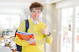 Young student man wearing headphones and backpack holding notebooks happy with big smile doing ok sign, thumb up with fingers,