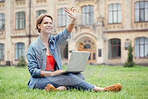 Young student at university campus sitting on grass using laptop waving to friend positive