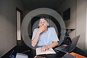 A young student yawns while reading a book. The student wants to sleep. Homework. Teaching at home.