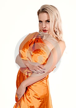 Young stunning tanned blonde woman with long curly hair wearing orange tunic dress posing at camera on a white