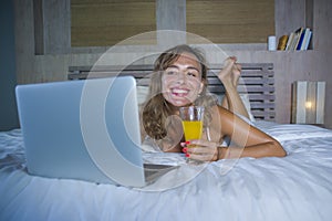 Beautiful Caucasian woman 30s lying on bed at night in home bedroom using internet at laptop computer drinking healthy orange juic