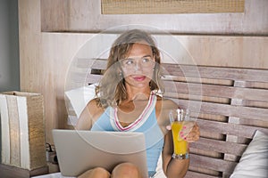 Beautiful Caucasian woman 30s sitting on bed at night in home bedroom using internet at laptop computer drinking healthy orange ju